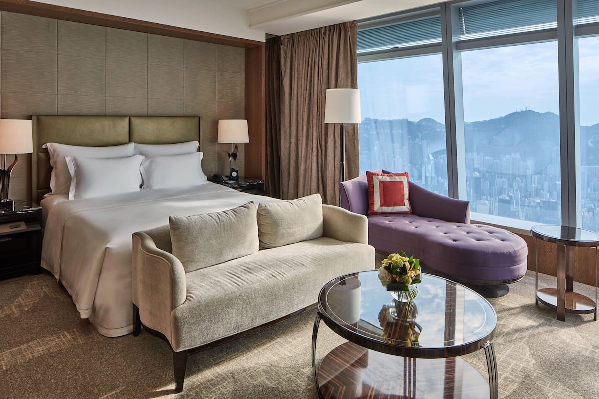 A hotel room with a large bed, a sofa, a round table, and a view of a cityscape through large windows.
