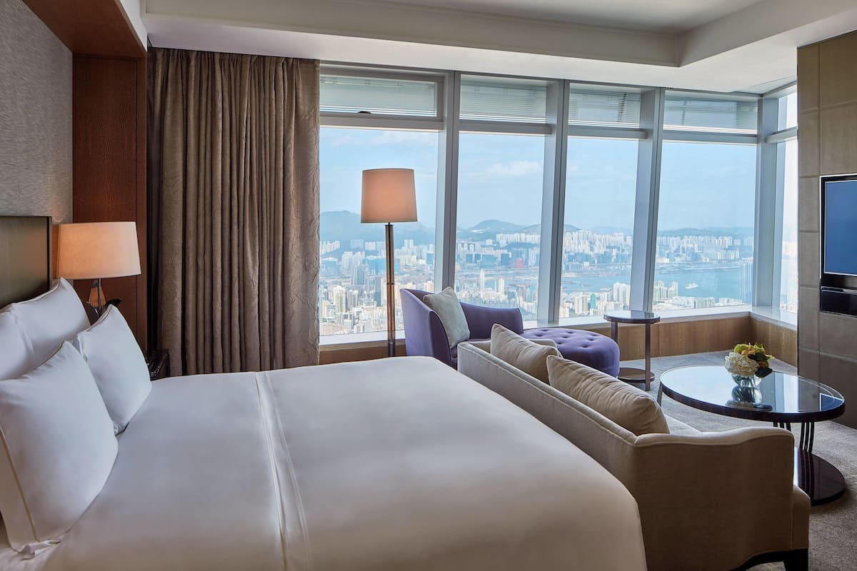 A hotel room with a large bed, a sofa, a small table, a lamp, and a TV. The room has large windows with a city view.