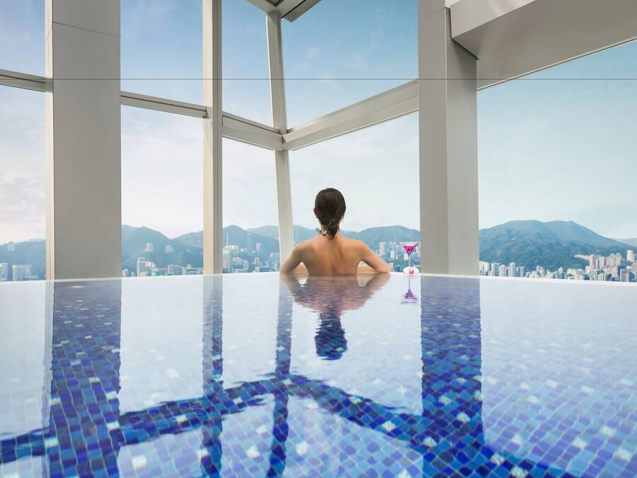 Un hombre descansando en una piscina infinita moderna, con un paisaje urbano y montañas al fondo.