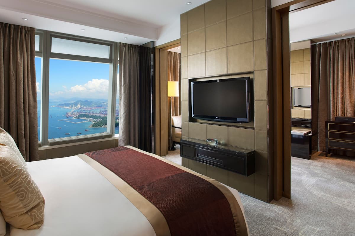 A hotel room with a large bed, a flat-screen TV mounted on the wall, a view of the sea and a bridge, and a desk with a lamp.
