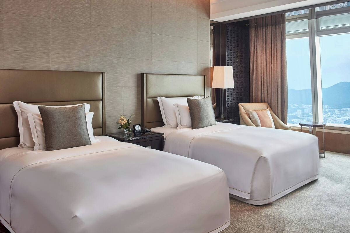 A hotel room with two beds, a nightstand, a lamp, and a view of the city.