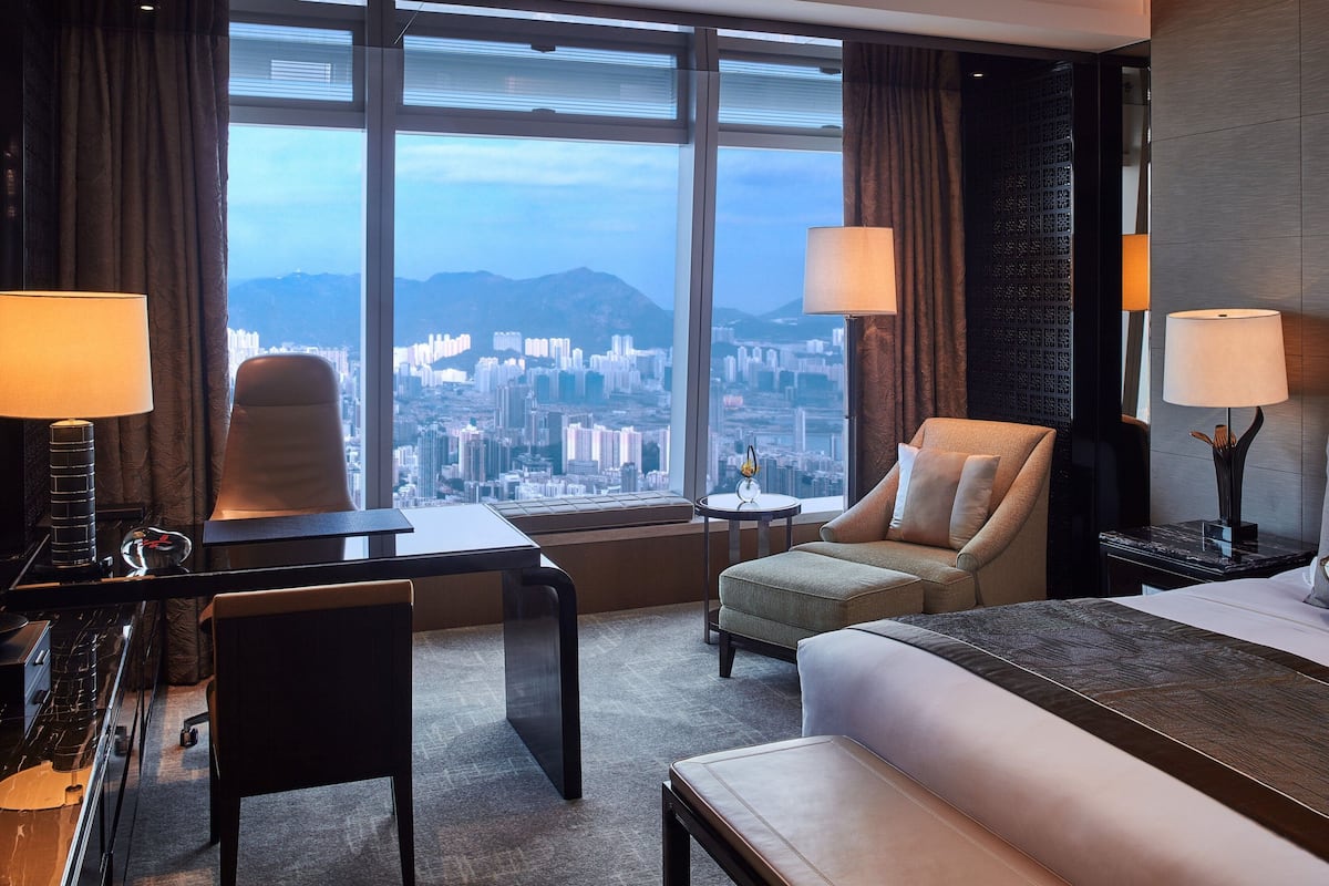 A hotel room with a large window offering a city view, a bed, a desk, a chair, and two lamps.