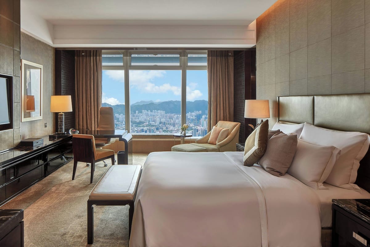 A hotel room with a large bed, a desk, a chair, and a view of a cityscape through large windows.