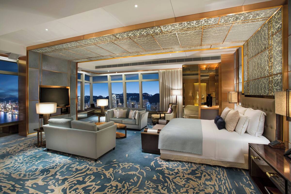 A spacious hotel room with a large bed, a sofa, a desk, and a television. The room has a city view and a patterned carpet.