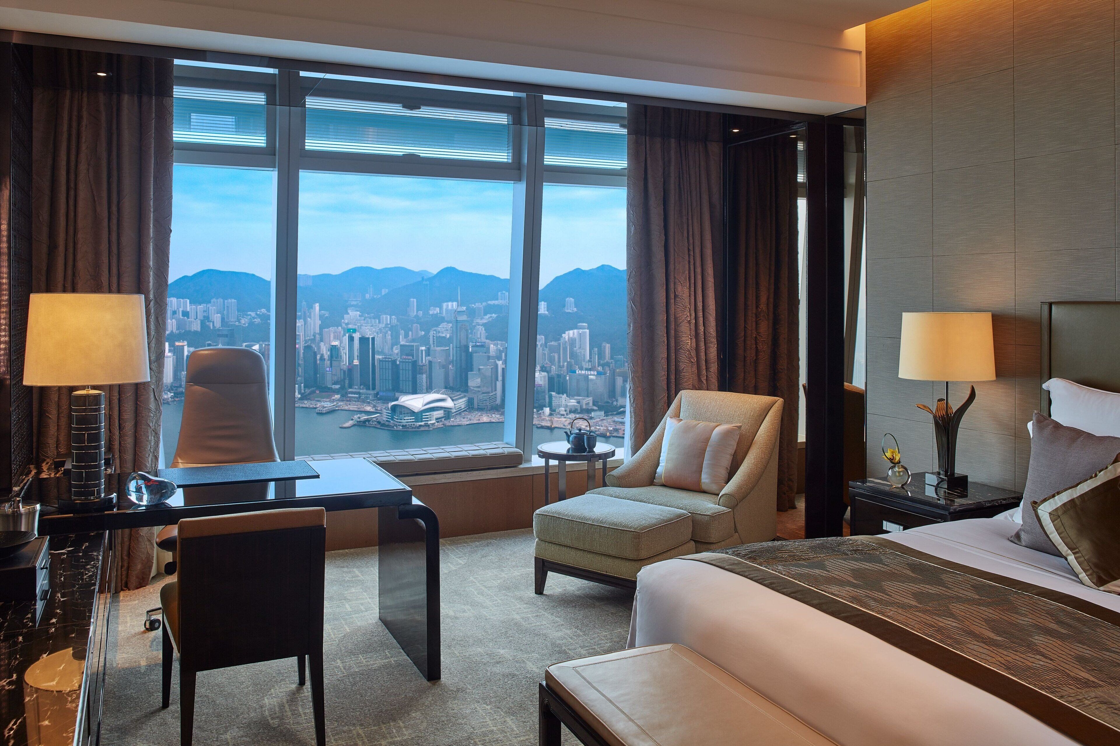 A hotel room with a large window, a desk, a chair, a bed, and a nightstand with a lamp.
