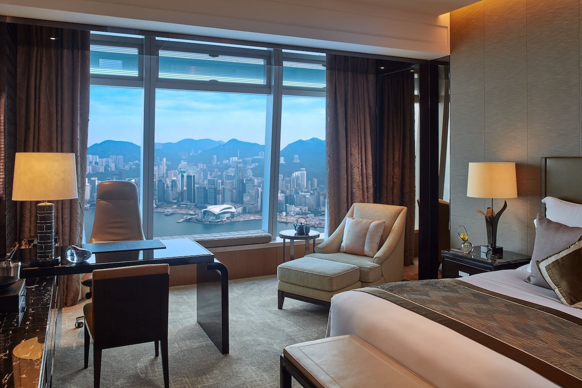 A hotel room with a large window, a desk, a chair, a bed, and a nightstand with a lamp.