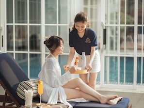 A person in a white robe sitting on a lounge chair by a pool, receiving a tray with amenities from a hotel staff member. - Hotel de l'Opera Hanoi - Mgallery (Hanoi)