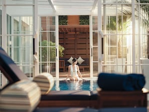 Ett poolområde med en solstol, en person som sitter i poolen och en träveranda med en soffa och växter.