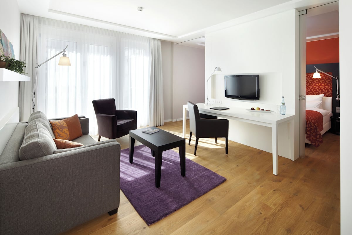Quarto de hotel moderno com sofá, mesa de jantar, televisão e cama com cabeceira vermelha.