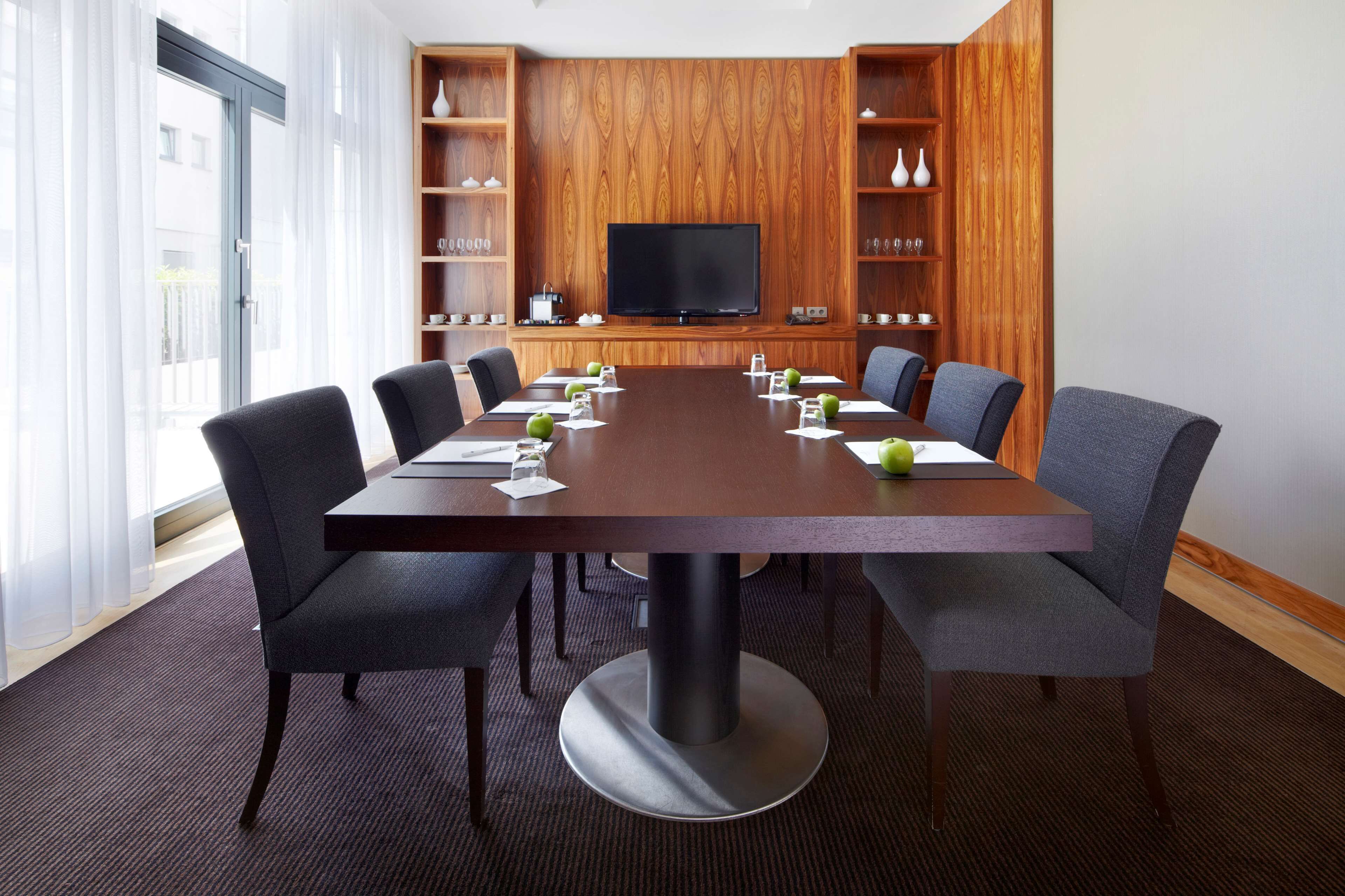A conference room with a large table, six chairs, a television, wooden shelves with decorative items, and a window with sheer curtains.