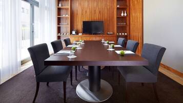 A conference room with a large table, six chairs, a television, wooden shelves with decorative items, and a window with sheer curtains.