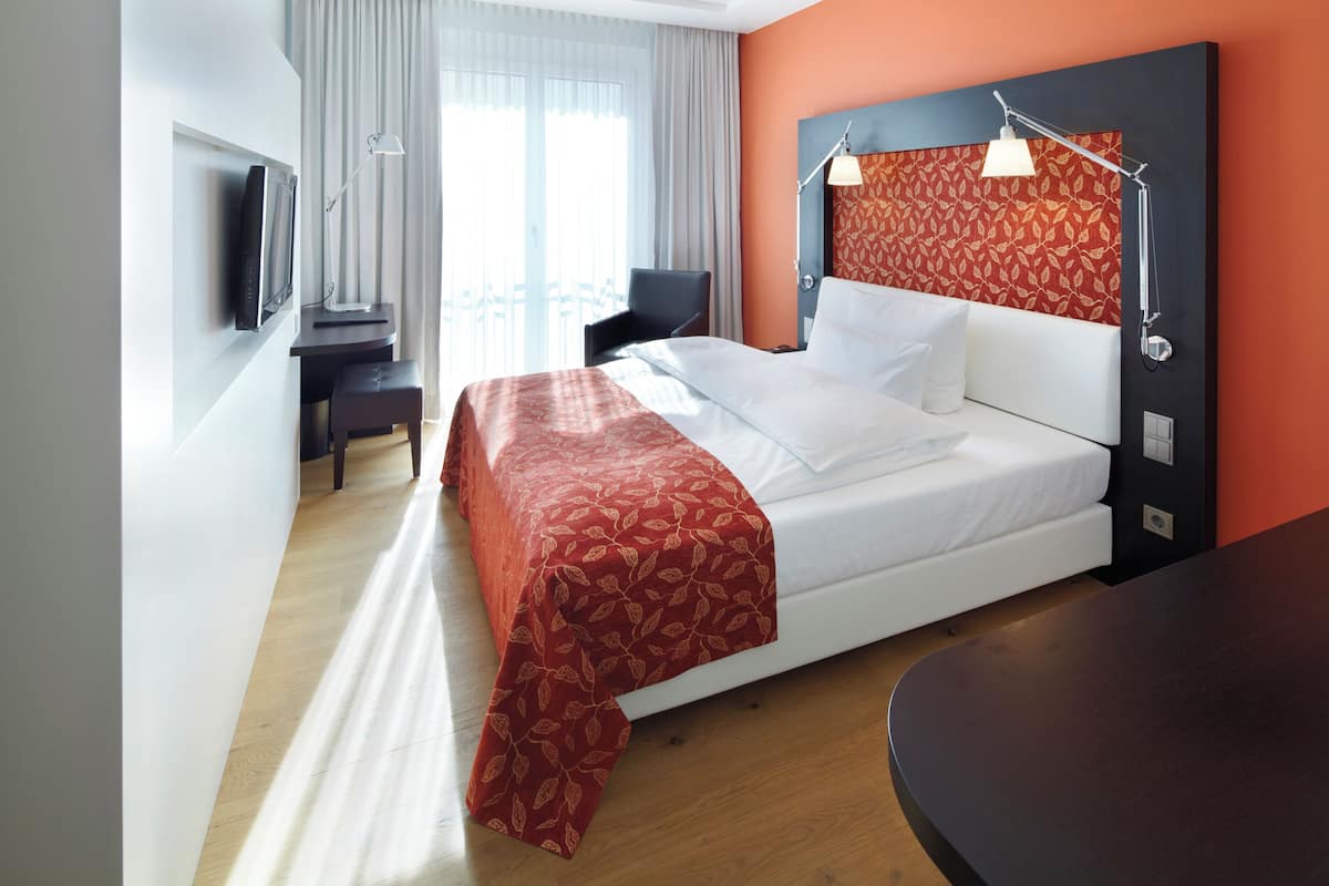 Quarto de hotel com cama, escrivaninha, cadeira, televisão e janela com cortinas.