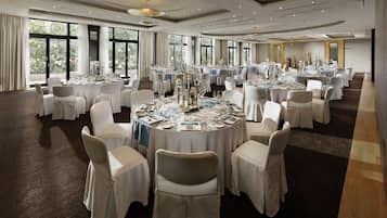 Un salón de banquetes con mesas redondas dispuestas para eventos, amplias ventanas y un techo alto con iluminación empotrada.