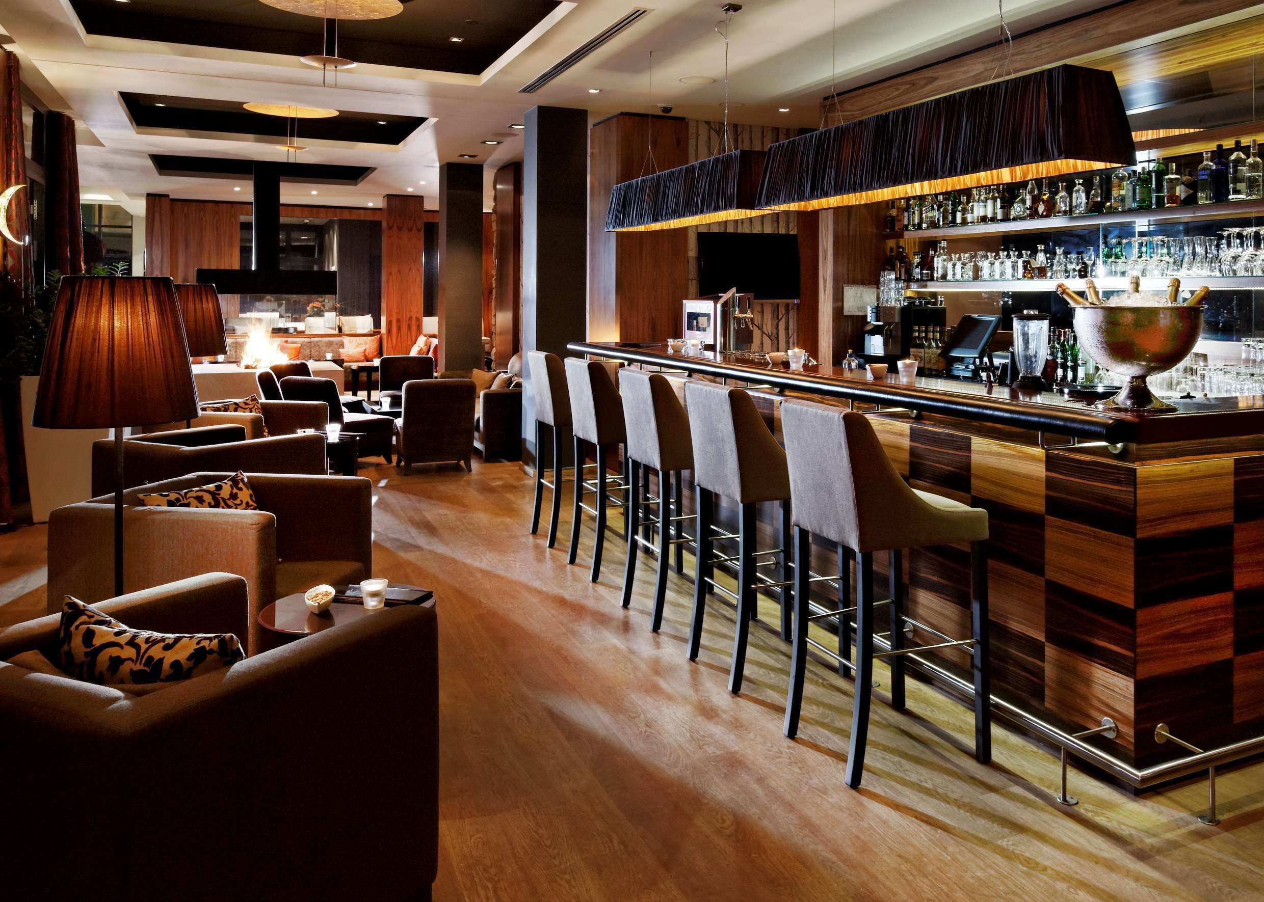 A bar area with wooden bar stools, a counter with bottles, and a seating area with sofas and lamps.