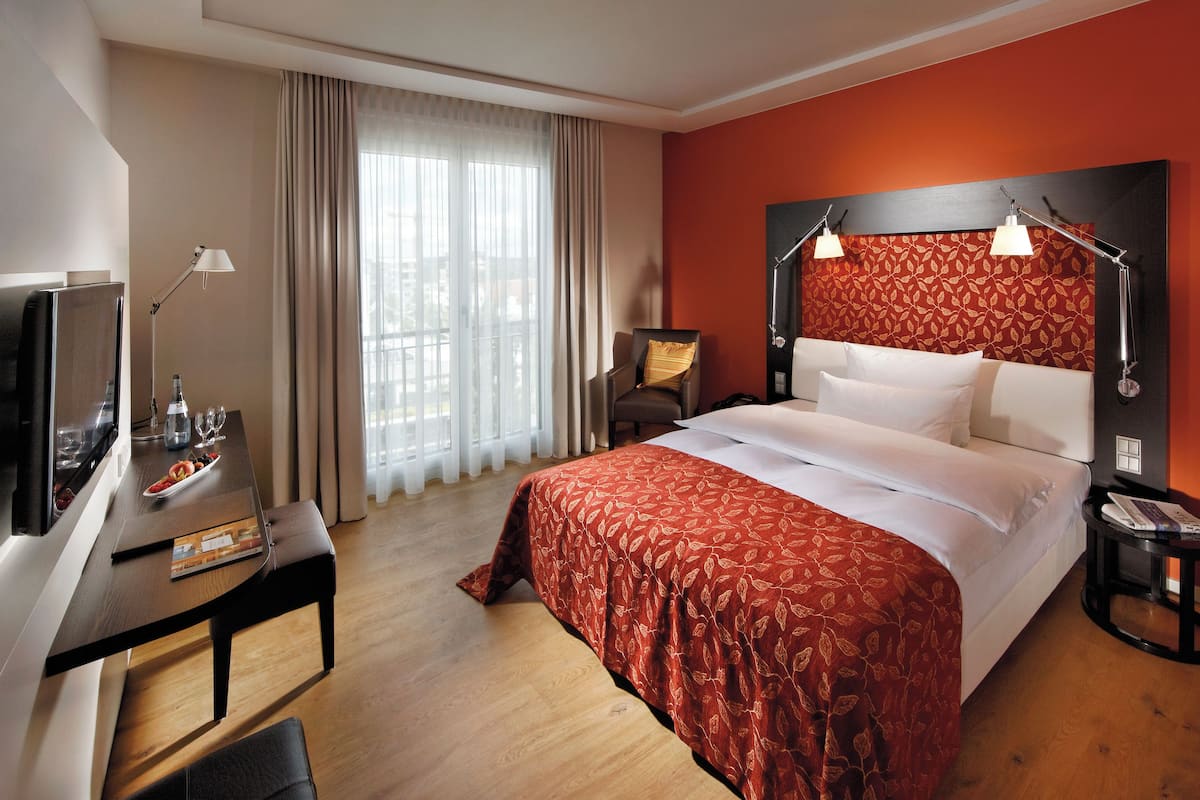 Quarto de hotel com cama, escrivaninha com luminária, televisão e janela com cortinas.