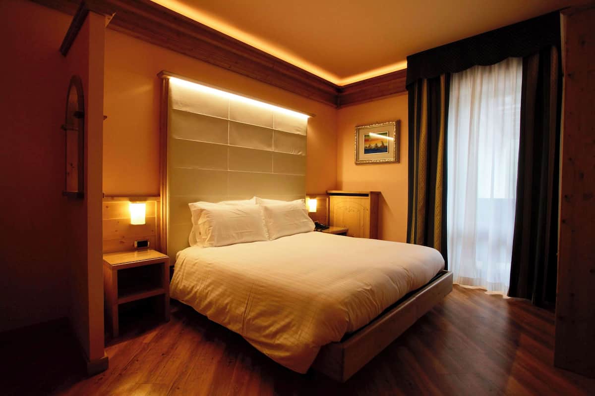Una habitación de hotel con una cama de madera, mesitas de noche, una lámpara de pared y un ventanal con cortinas.