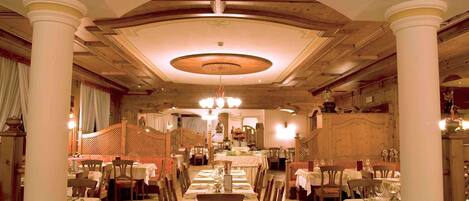 A formal dining room with multiple tables and chairs, a chandelier, and classical architectural details like columns and a decorative ceiling.