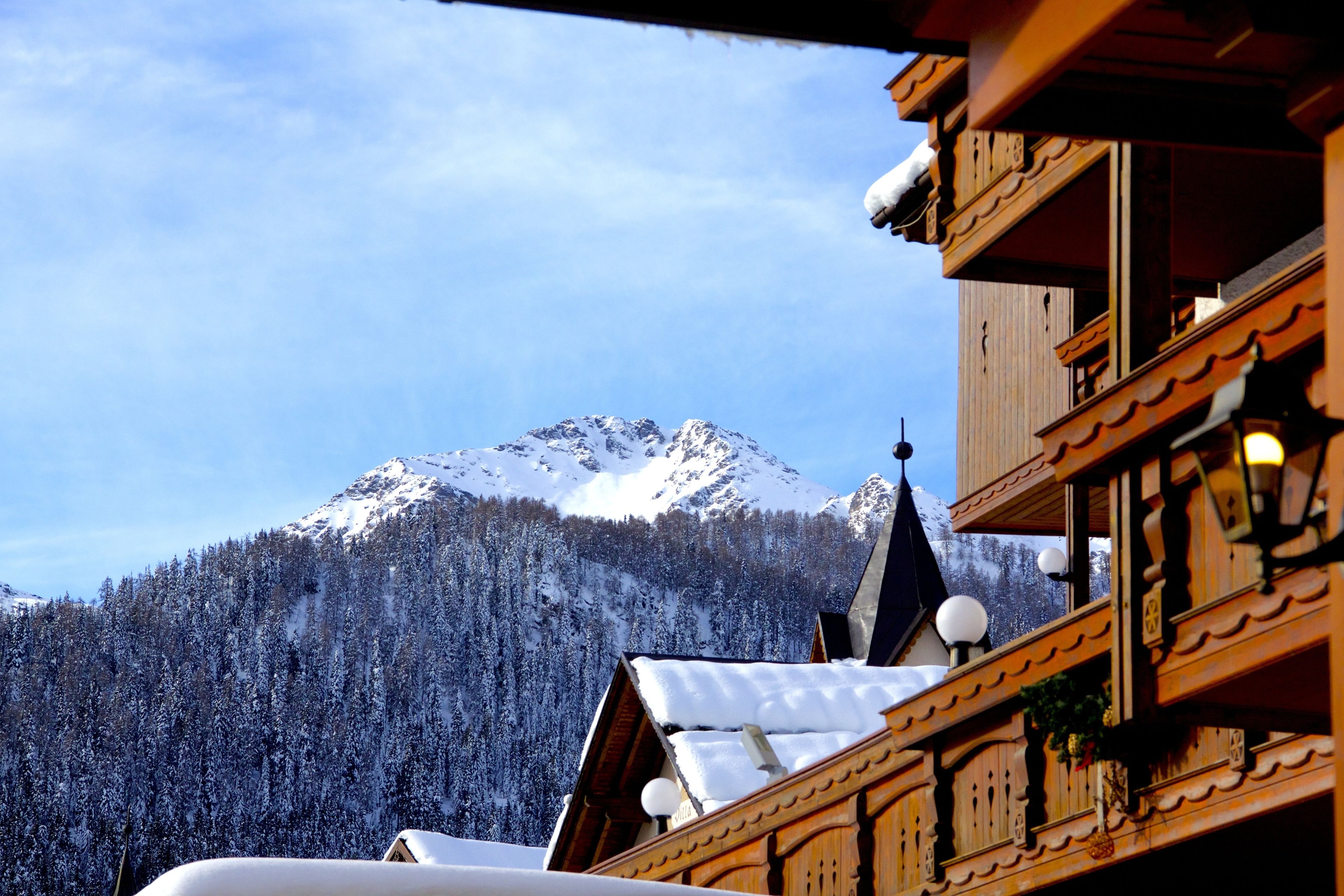 A wooden chalet with a snow-covered roof, a mountain backdrop, and a forested valley.
