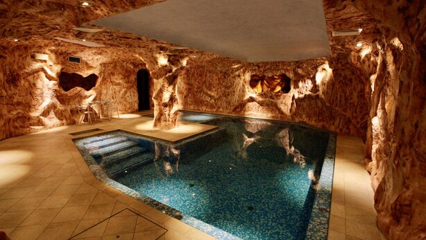 An indoor swimming pool with a natural rock wall design, a clear water surface, and a tiled floor.