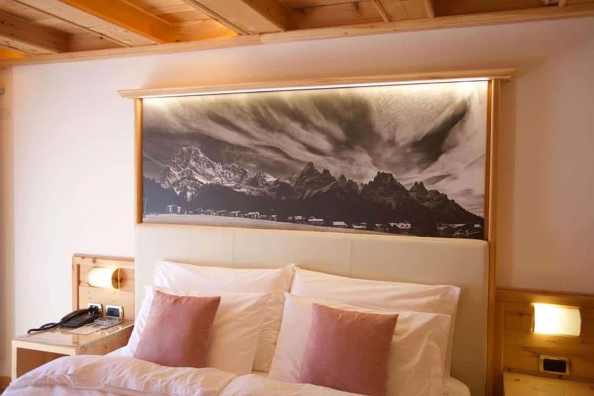 Una habitación de hotel con una cama, dos mesitas de noche con lámparas y un cuadro grande con un paisaje de montañas en la pared.