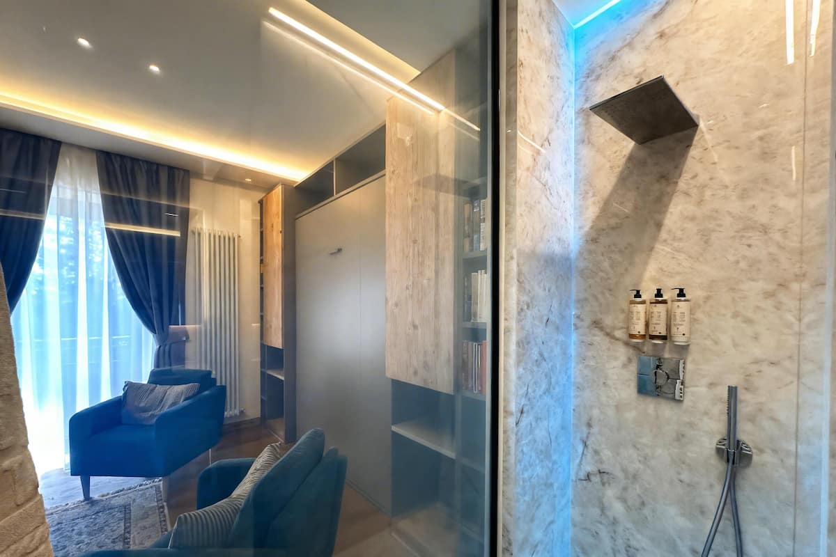 Un baño moderno con cabina de ducha de vidrio, paredes de mármol y una zona de estar con una silla y cojines azules.