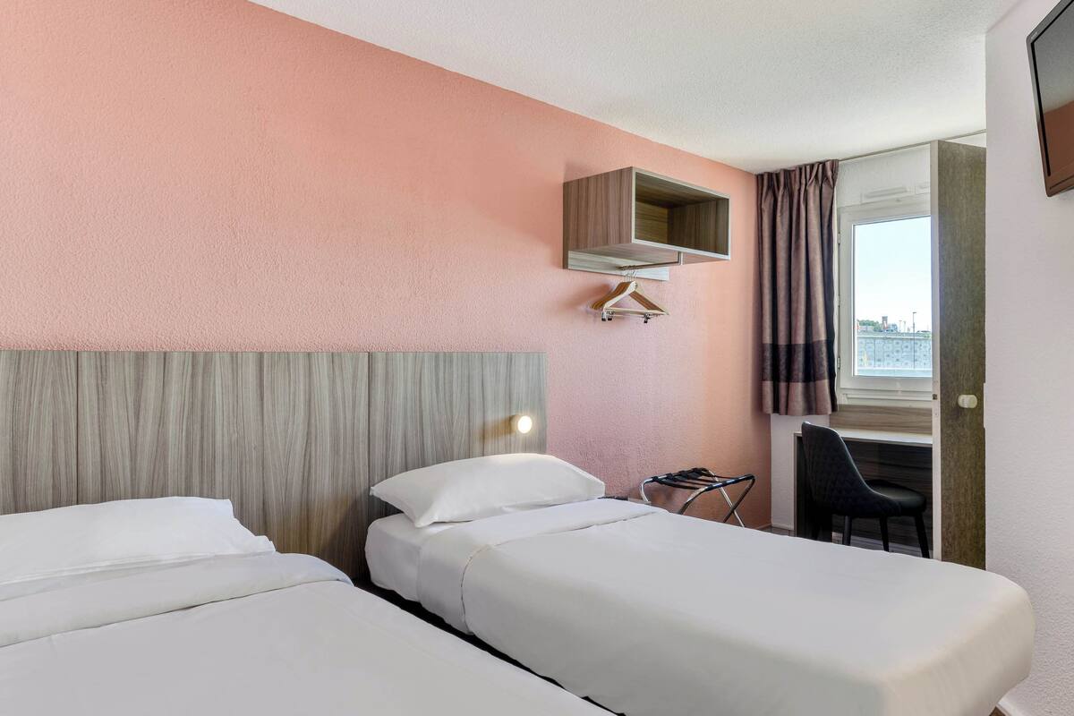 A hotel room with two beds, a desk, a chair, a hanging coat rack, and a window with curtains.