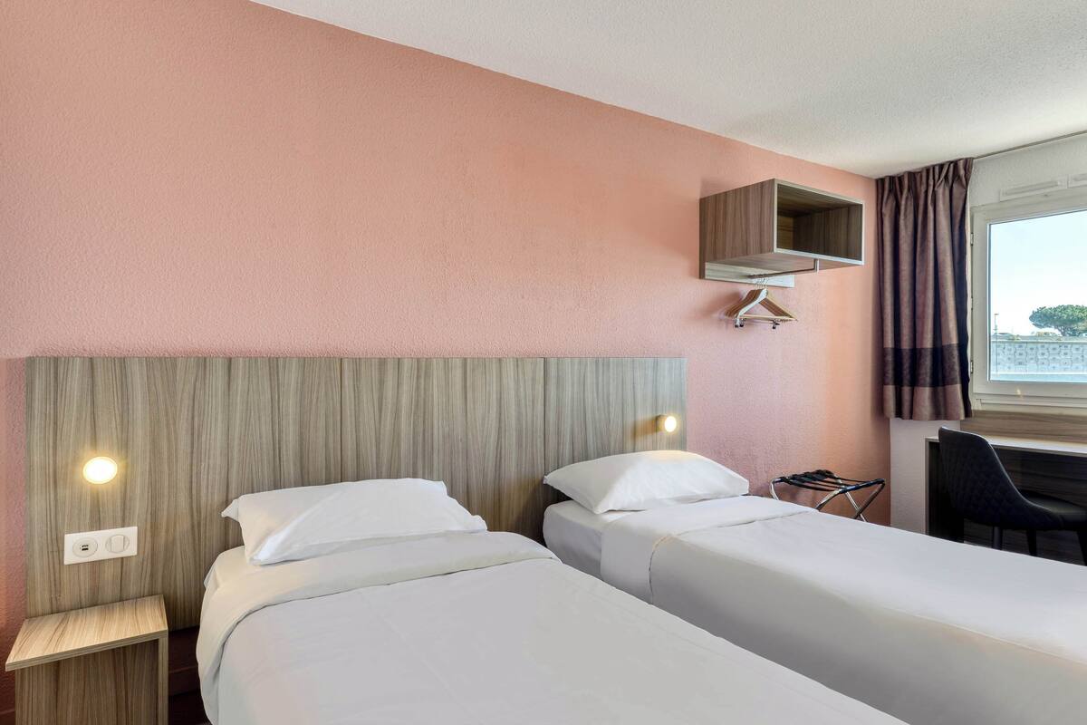 A hotel room with two single beds, a wooden headboard, a small desk, and a window with curtains.