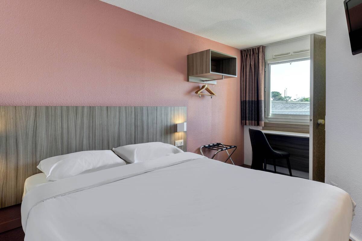 A hotel room with a bed, a desk, a hanging coat rack, and a window with curtains.