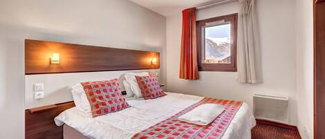 A hotel room with a large bed, two bedside tables, a headboard with wall-mounted lights, a window with curtains, and a radiator.