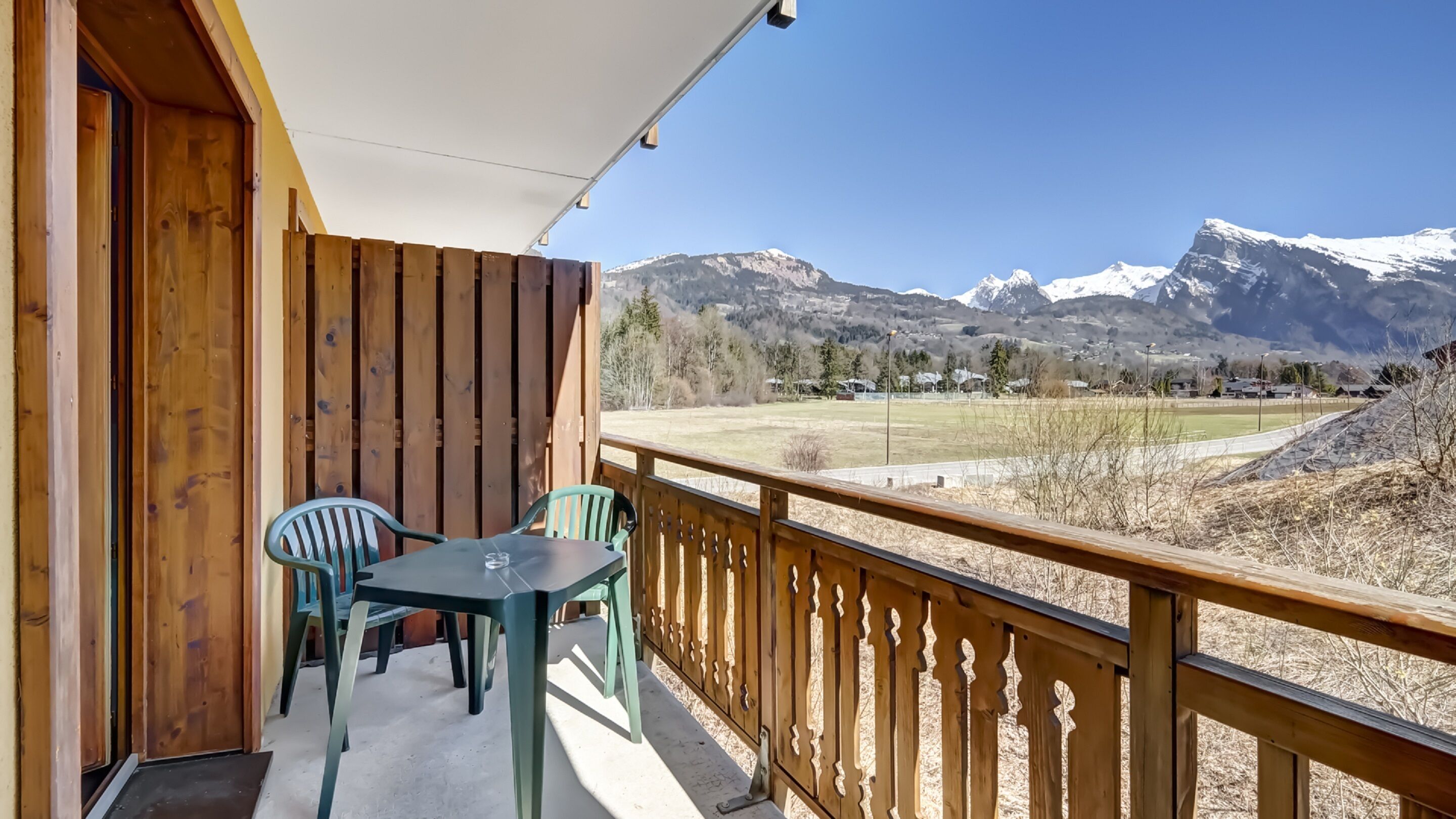 A balcony with wooden railings, a table, and two chairs. It overlooks a mountainous landscape with snow-capped peaks and a clear sky.