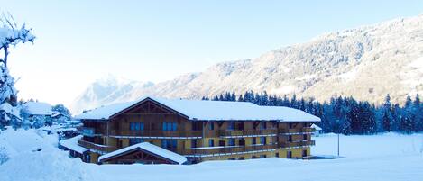 A multi-story lodge with a snow-covered roof and balconies, surrounded by snow-covered trees and mountains.