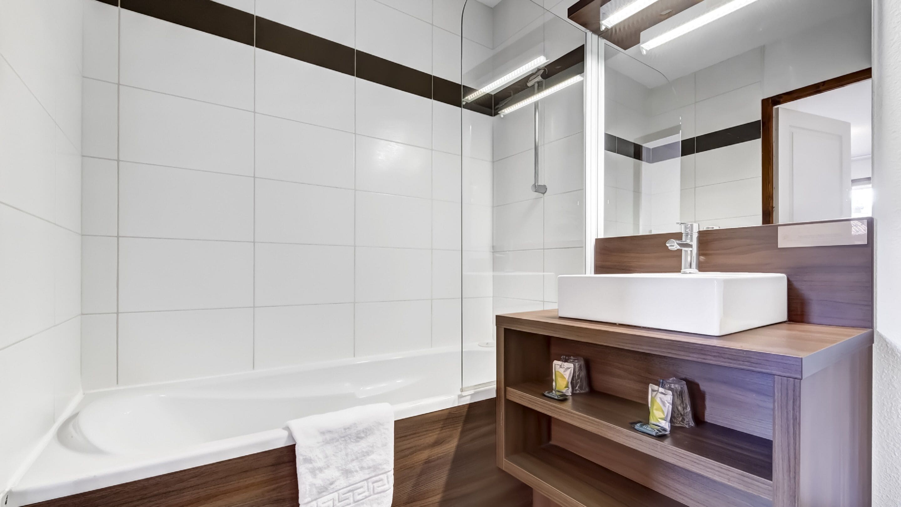 A modern bathroom with white tiles, a wooden vanity, a large mirror, and a bathtub.