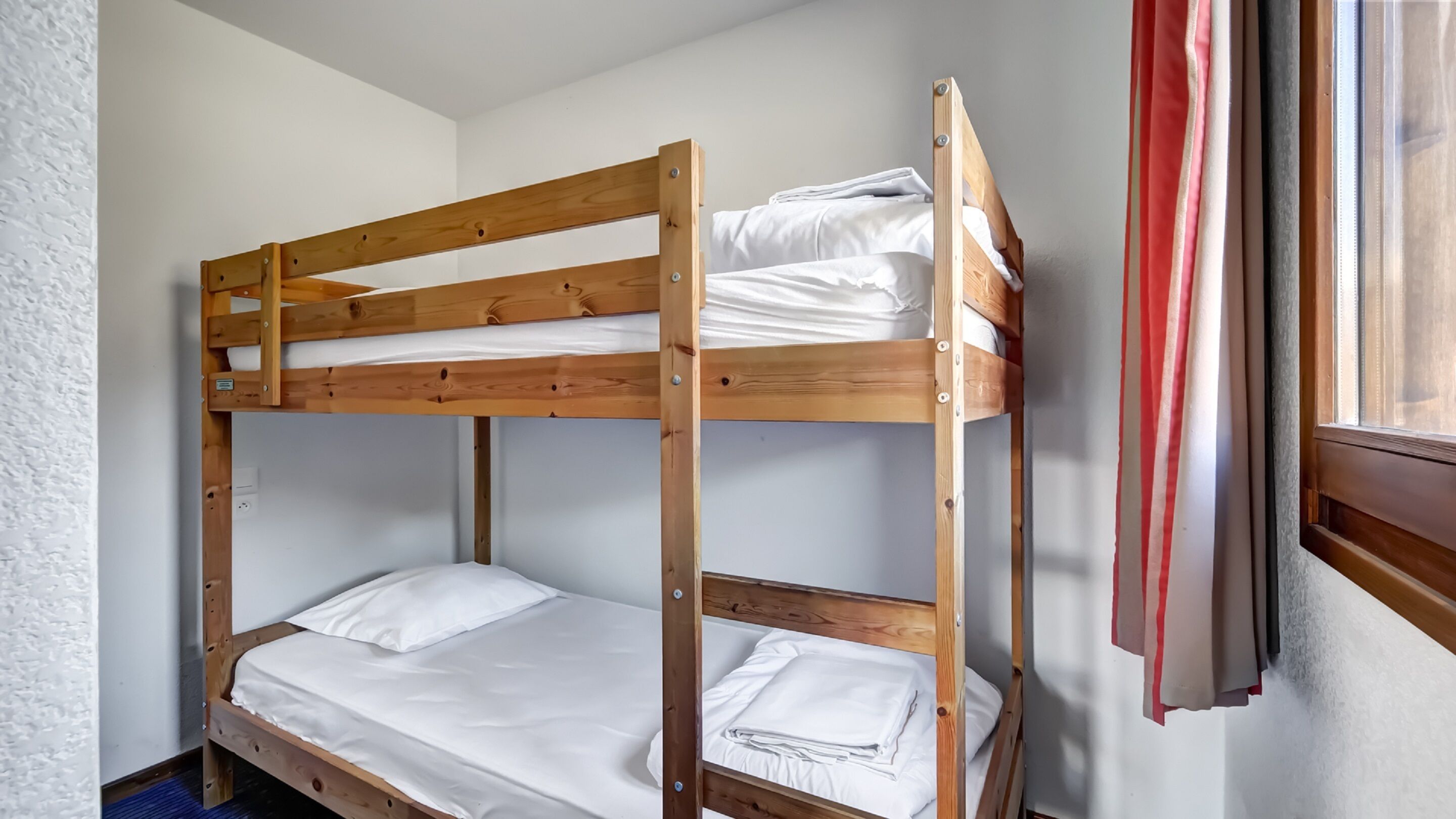 Una cama litera con ropa blanca y una ventana con cortinas rojas.