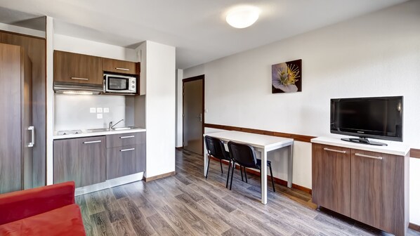 A compact kitchen with a microwave, sink, and cabinets, a dining area with chairs, and a TV on a cabinet. - Résidence Grand Massif - Vacancéole (Morillon)