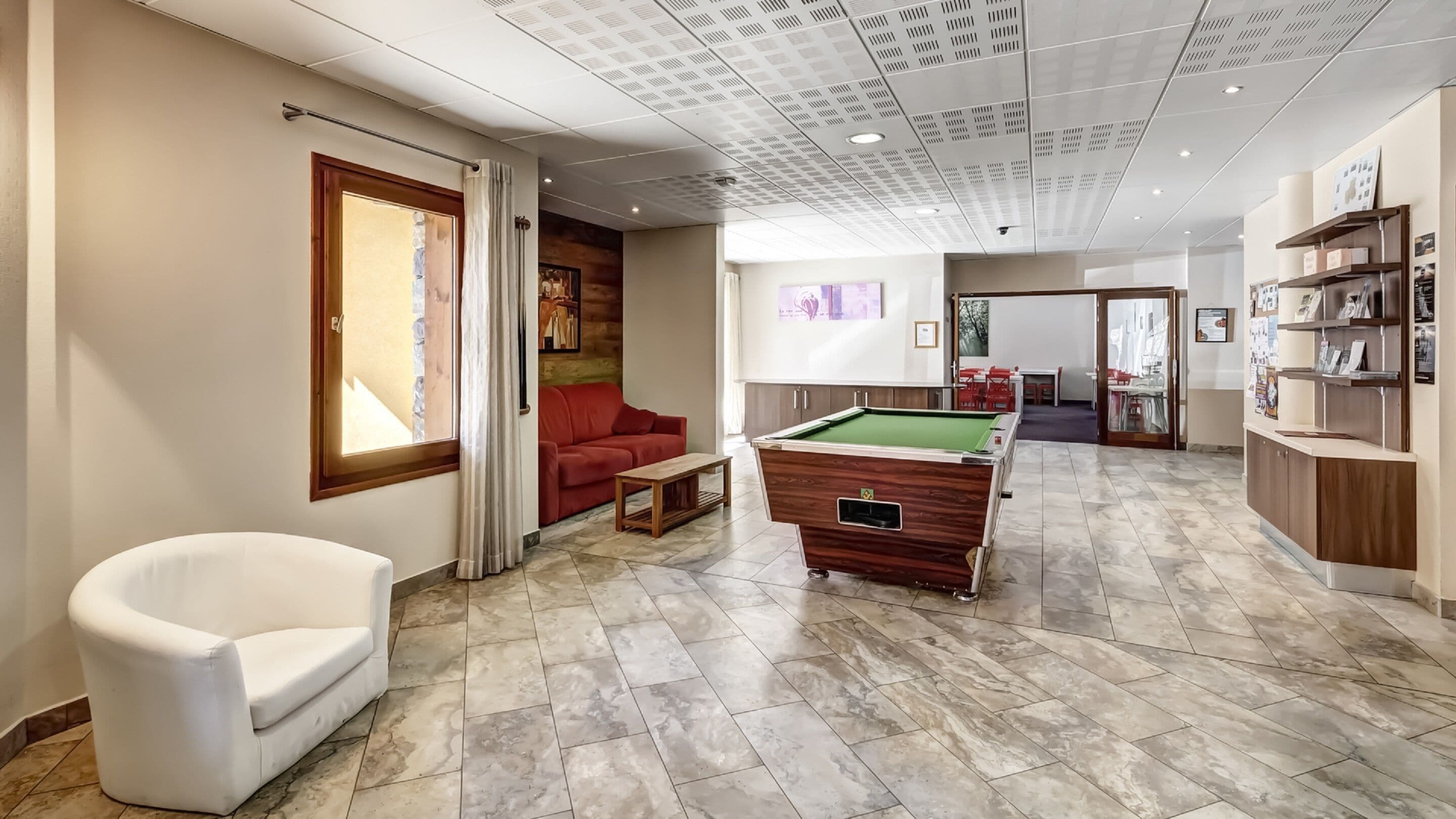 Une salle de billard équipée d’une table de billard, d’un canapé rouge, d’un fauteuil blanc et d’un comptoir d’accueil orné de tableaux encadrés.