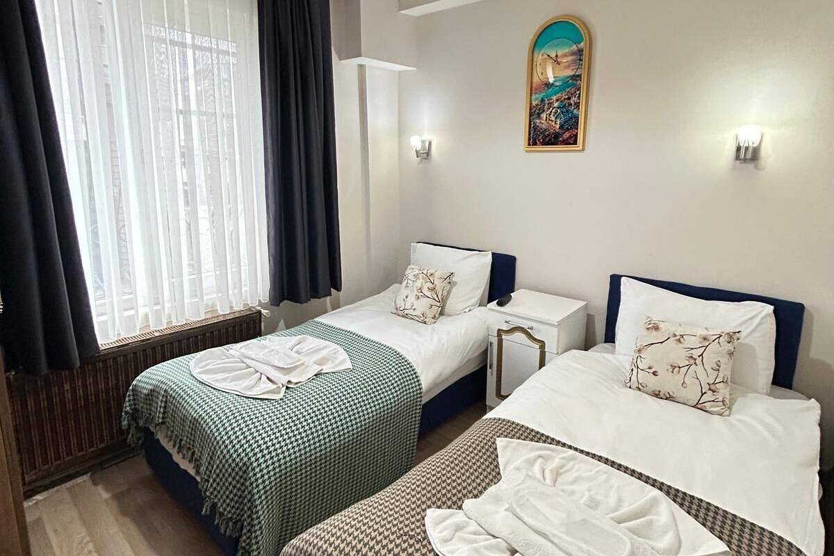 Ein Hotelzimmer mit zwei Betten, einem großen Fenster mit Vorhängen, einer Deckenleuchte, einem Wandbild und einem Nachttisch mit Lampe.