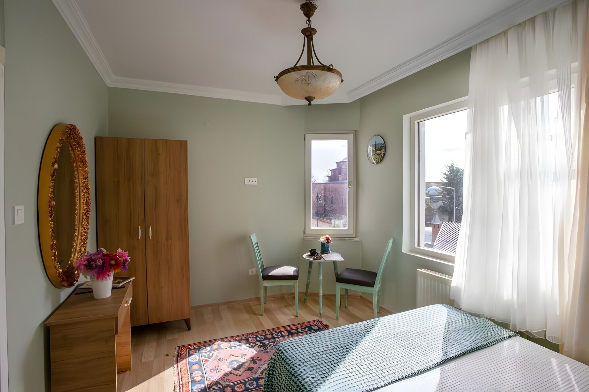 Een slaapkamer met een bed, een houten kledingkast, een bureau met spiegel en bloemen, twee stoelen, een kleine tafel, en een raam met gordijnen.
