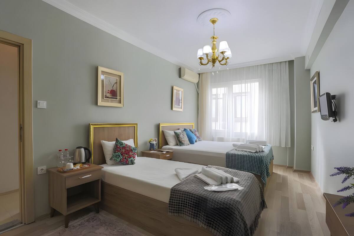Ein Hotelzimmer mit zwei Betten, einer Chandelüster, gerahmten Bildern, einem Fernseher und einem Fenster mit Vorhängen.