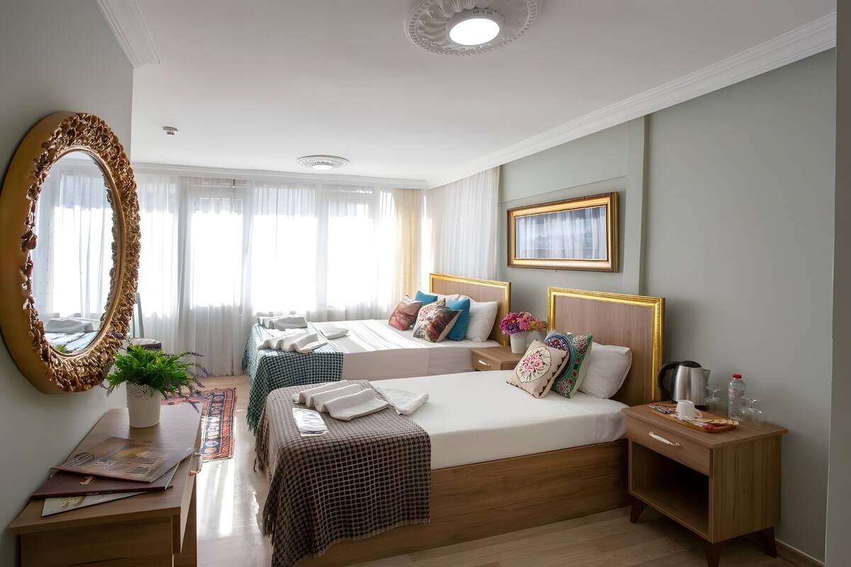 Ein Hotelzimmer mit zwei Betten, einem großen Spiegel, einem Nachttisch mit Pflanze und einem kleinen Tisch mit Wasserkocher und Gläsern.