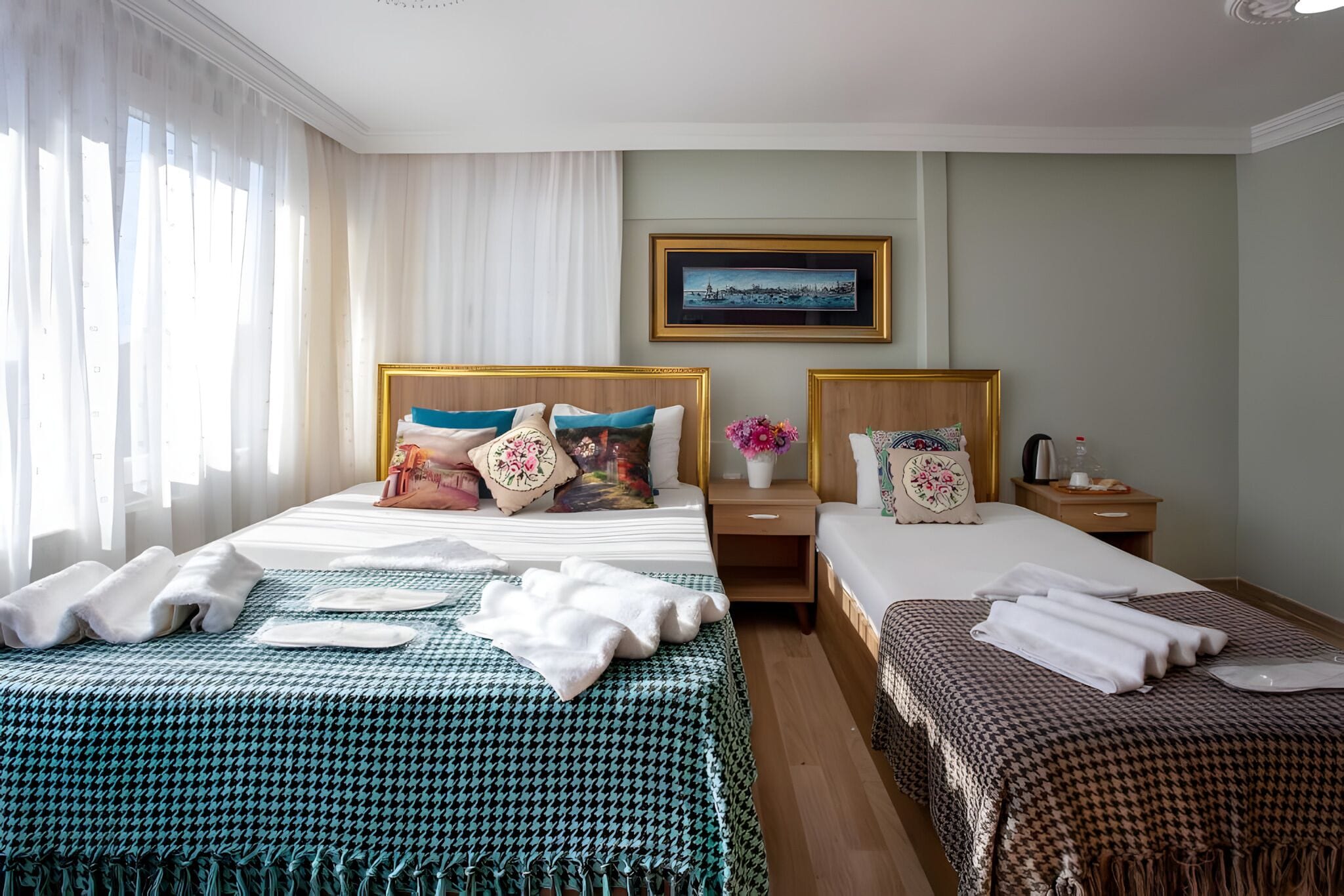 A hotel room with two beds, a large window with sheer curtains, a framed picture on the wall, and a bedside table with a vase of flowers.
