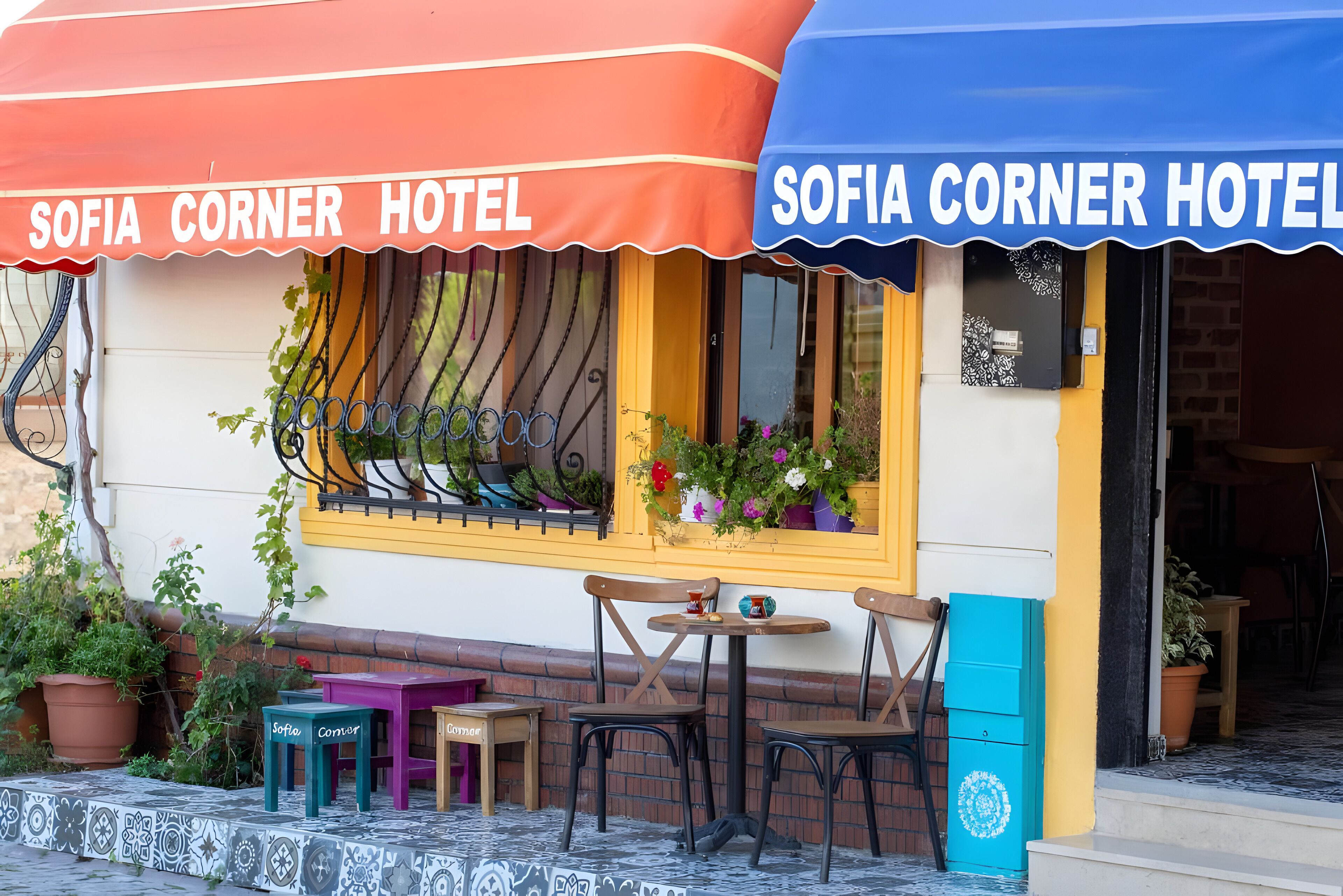 The exterior of Sofia Corner Hotel with a colorful awning, outdoor seating, and potted plants.