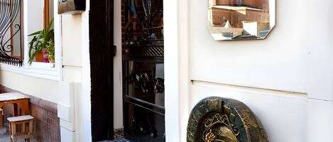 A building entrance with a tiled floor, a black ornate door, a small outdoor seating area, and a wall-mounted mirror reflecting a building.
