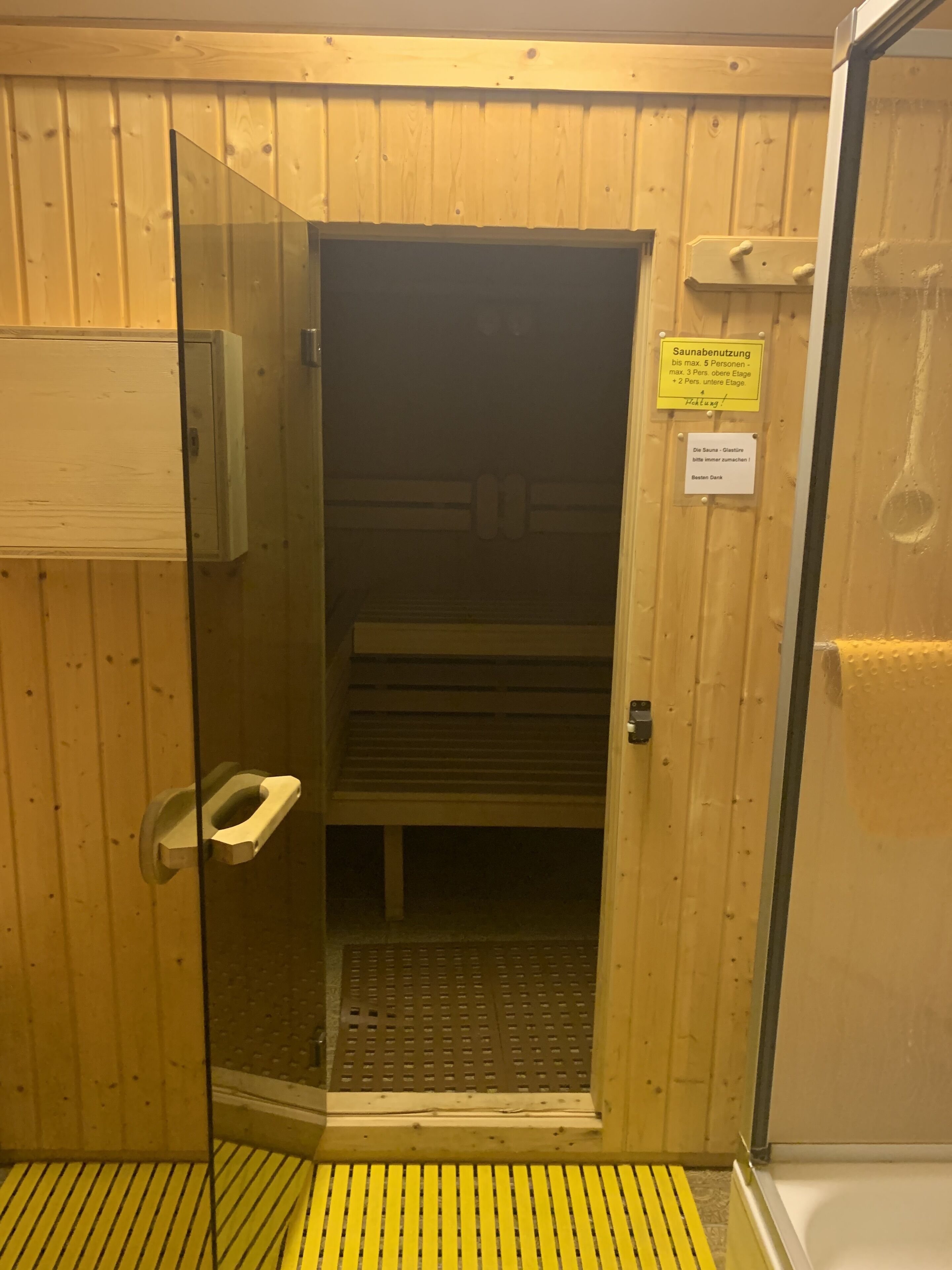 Un sauna avec un intérieur en bois, une porte en verre et un tapis antidérapant jaune à l’entrée.