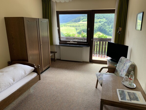 Een kamer met een bed, een bureau met een laptop, een televisie en uitzicht op een groen landschap door een raam.