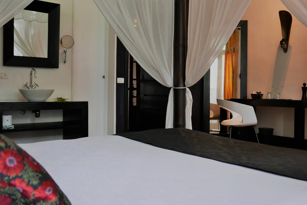 A modern hotel room with a large bed, a bedside table with a white bowl, a mirror, and a chair.