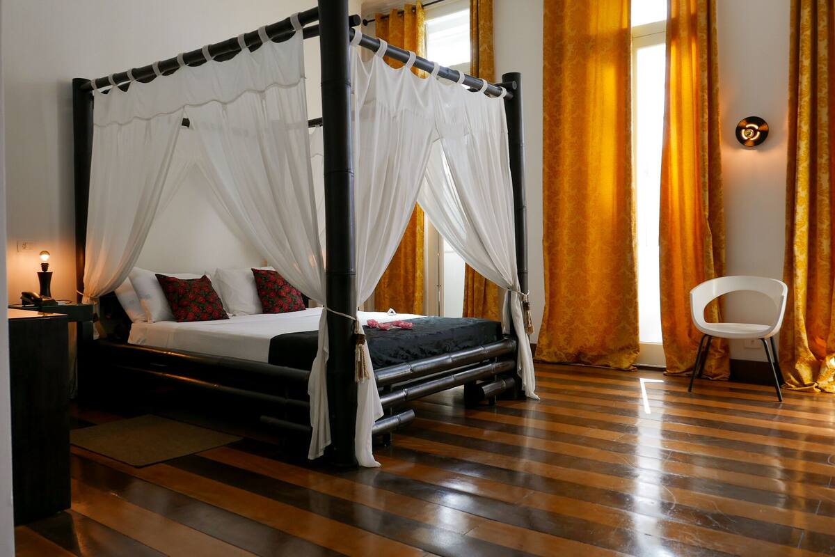 A four-poster bed with white drapes, a wooden floor, and a white chair.