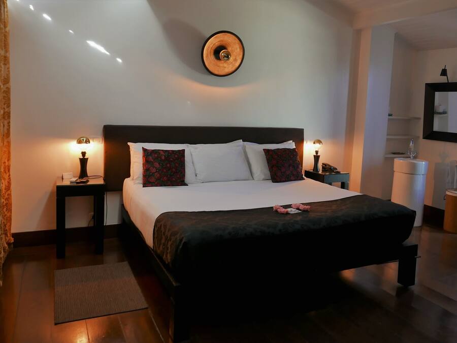 Uma cama bem arrumada com almofadas brancas e vermelhas, uma cabeceira escura e duas mesas de cabeceira com abajures.