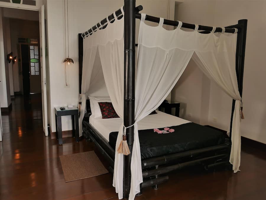 Un lit à baldaquin, une table de chevet avec une lampe et un banc dans une chambre d’hôtel.