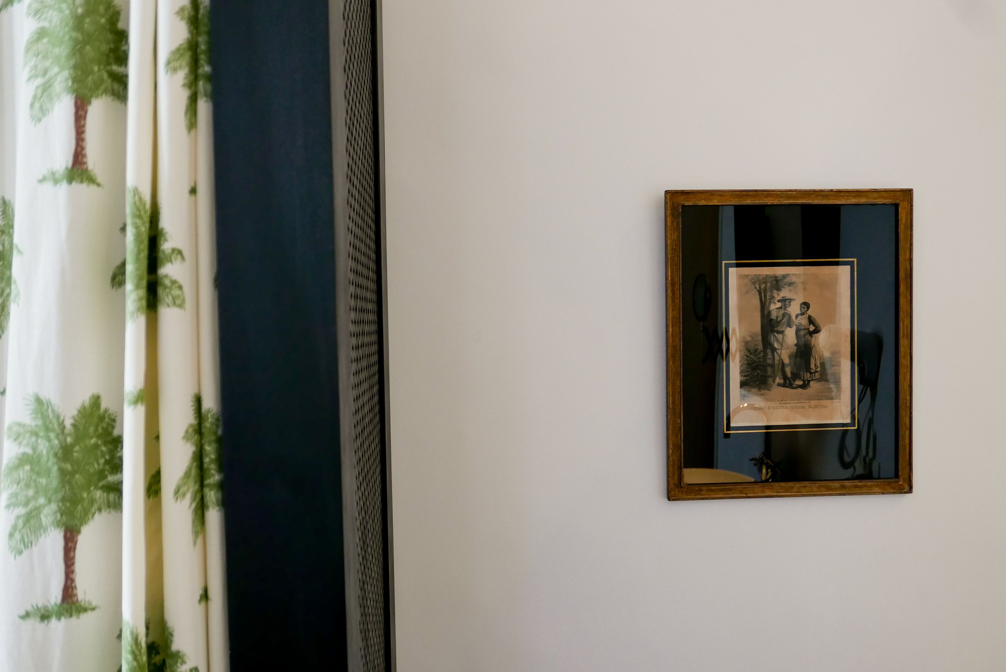 A framed picture on a wall, a window with a view of palm trees, and a curtain with a palm tree pattern.