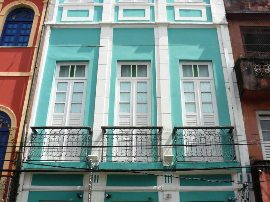 Un edificio de varios pisos con fachada azul verdosa y blanca, balcones con barandillas de hierro forjado y una puerta visible con placa de número.